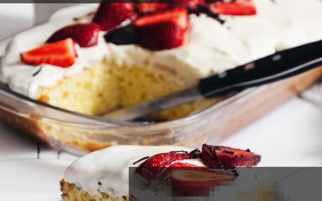 کیک سه شیر برش خورده با بافت اسفنجی و خامه سفید، تزئین شده با توت فرنگی تازه و پودر پسته روی میز چوبی